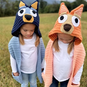 以下が含まれることがあります： 手作りのかぎ針編みの帽子とショールをかぶった2人の子供たち。1つの帽子はネイビーブルーに黄色とタンのアクセント、もう1つはオレンジにタンと白のアクセント。ショールは水色とピーチ色で、ポケットが付いています。