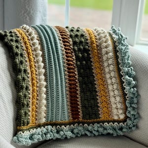 Puede incluir: Una manta de ganchillo hecha a mano con una variedad de puntadas texturizadas y una paleta de colores verde oliva, amarillo mostaza, marrón y crema. La manta tiene un borde festoneado azul claro y está colocada sobre una silla gris.