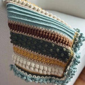 May include: A hand-crocheted blanket with a striped design in green, blue, brown, yellow, and cream. The blanket has textured bobble stitches and a scalloped edge. The blanket is draped over a white surface.