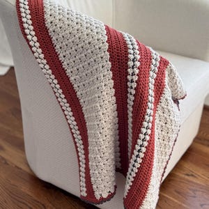 Puede incluir: Una manta de ganchillo hecha a mano sobre un sillón blanco. La manta presenta rayas alternas de hilo beige y color óxido. Los bordes están acabados con un borde gris oscuro, creando un aspecto acogedor y texturizado. La manta es un artículo de decoración suave.