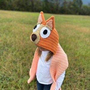 K&ouml;nnte beinhalten: Ein orangefarbener H&auml;kelhut mit hunde&auml;hnlichen Ohren und einem passenden pfirsichfarbenen Schal. Der Hut hat eine braune Nase, wei&szlig;e Augen und schwarze Pupillen. Der Schal hat Taschen und ist &uuml;ber einer Person drapiert, die ein wei&szlig;es Hemd und blaue Jeans tr&auml;gt.