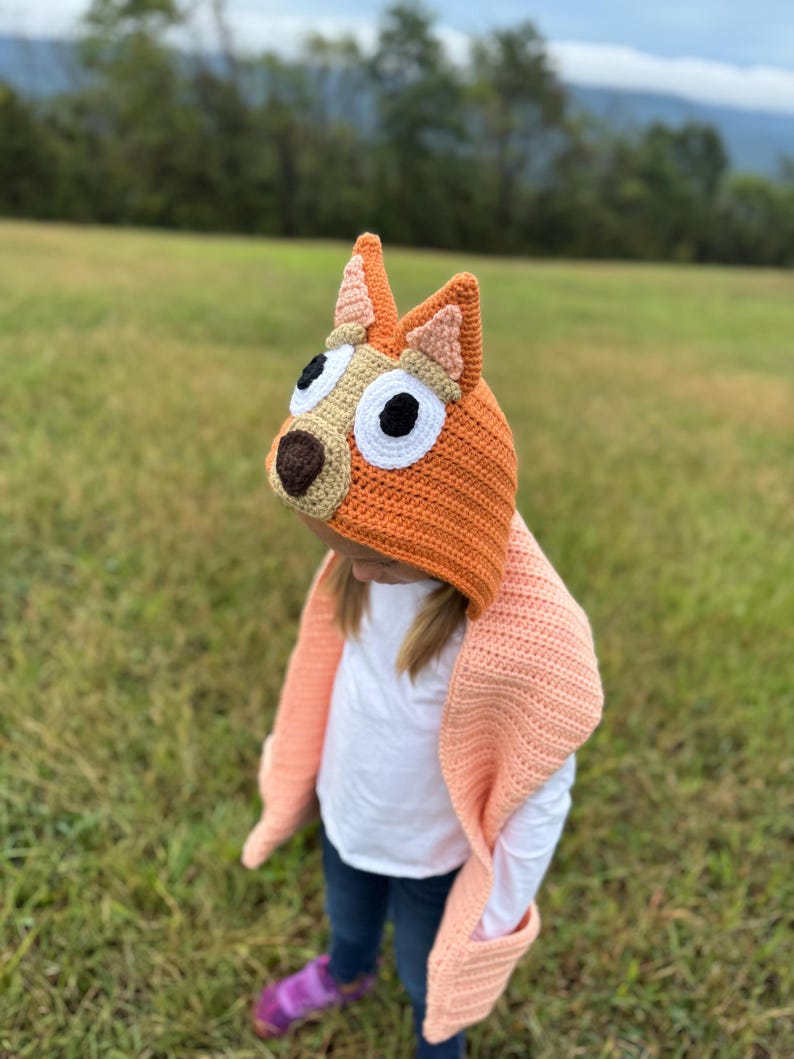 K&ouml;nnte beinhalten: Ein orangefarbener H&auml;kelhut mit hunde&auml;hnlichen Ohren und einem passenden pfirsichfarbenen Schal. Der Hut hat eine braune Nase, wei&szlig;e Augen und schwarze Pupillen. Der Schal hat Taschen und ist &uuml;ber einer Person drapiert, die ein wei&szlig;es Hemd und blaue Jeans tr&auml;gt.