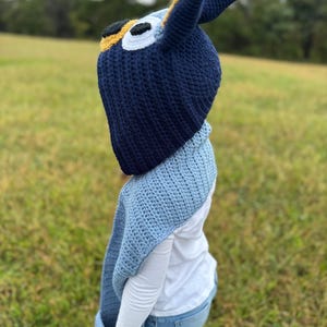 K&ouml;nnte beinhalten: Eine marineblaue H&auml;kelm&uuml;tze mit gelben und wei&szlig;en Akzenten, die einer Zeichentrickfigur &auml;hnelt. Die M&uuml;tze hat gro&szlig;e, spitze Ohren. Ein hellblauer H&auml;kelschal ist ebenfalls zu sehen.