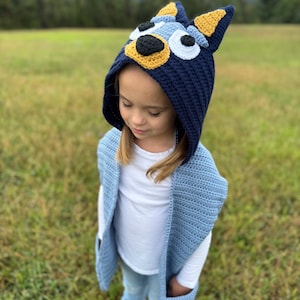K&ouml;nnte beinhalten: Ein Kind tr&auml;gt eine handgefertigte, geh&auml;kelte Bluey-inspirierte Kapuzenm&uuml;tze und eine passende blaue Weste mit Taschen. Die M&uuml;tze hat eine dunkelblaue Basis mit gelben und hellblauen Akzenten, die der Zeichentrickfigur &auml;hneln. Das Kind tr&auml;gt ein wei&szlig;es Hemd und blaue Jeans.