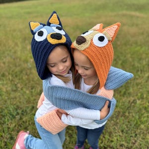 K&ouml;nnte beinhalten: Zwei Kinder tragen handgefertigte Hunde-H&auml;kelm&uuml;tzen, eine marineblaue und eine orangefarbene. Sie umarmen sich, mit blauen und pfirsichfarbenen H&auml;kelschal um sich. Die M&uuml;tzen zeigen detaillierte Hundegesichter.