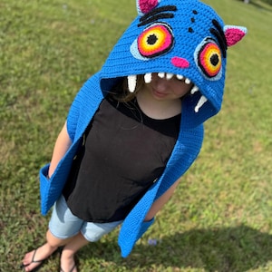May include: A blue crochet hooded cowl featuring a creature design with large yellow and orange eyes, black eyebrows, pink inner ears, and white pointed teeth along the hood opening. The garment includes long side panels with integrated pockets.