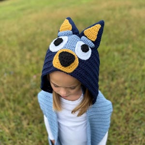 K&ouml;nnte beinhalten: Eine marineblaue H&auml;kelm&uuml;tze mit einem Hunde-Gesicht-Design, mit gelben Ohren, wei&szlig;en Augen und einer schwarzen Nase. Die M&uuml;tze wird von einer Person getragen, die eine hellblaue H&auml;kelweste tr&auml;gt.