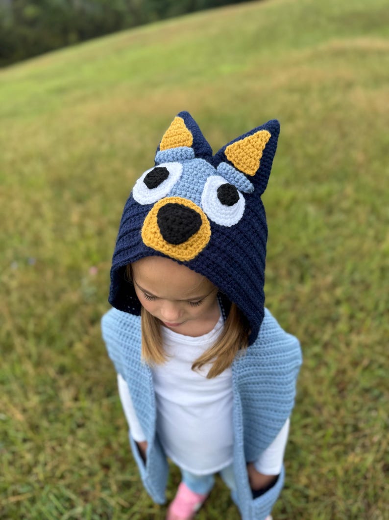 K&ouml;nnte beinhalten: Eine marineblaue H&auml;kelm&uuml;tze mit einem Hunde-Gesicht-Design, mit gelben Ohren, wei&szlig;en Augen und einer schwarzen Nase. Die M&uuml;tze wird von einer Person getragen, die eine hellblaue H&auml;kelweste tr&auml;gt.