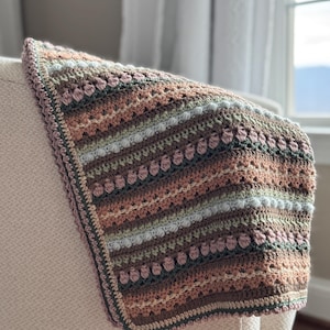 Puede incluir: Una manta de ganchillo con un patrón a rayas en tonos de marrón, verde, rosa y blanco. La manta tiene un borde de ganchillo en un tono de marrón más oscuro.