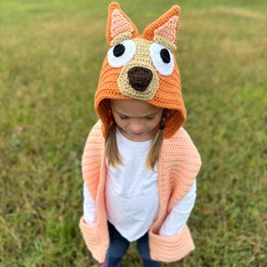 K&ouml;nnte beinhalten: Ein geh&auml;kelter orangefarbener Hundehut mit gro&szlig;en wei&szlig;en Augen, einer braunen Nase und beigefarbenen Ohren. Der Hut wird von einer Person getragen, die eine pfirsichfarbene geh&auml;kelte Weste und lila Stiefel tr&auml;gt. Der Hintergrund ist ein grasbewachsenes Feld.