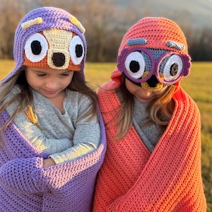 May include: Two children wearing handmade crochet owl hats and matching wraps. The hats feature large, expressive eyes, beaks, and ear details. One hat is purple and orange, the other is pink and purple. The wraps are crocheted in coordinating colours.