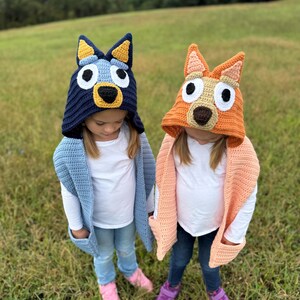 K&ouml;nnte beinhalten: Zwei Kinder tragen handgefertigte H&auml;kelh&uuml;te und -schals. Ein Hut ist marineblau mit gelben Akzenten, der einem Zeichentrickhund &auml;hnelt. Der andere ist orange mit beigen Akzenten. Beide Kinder tragen wei&szlig;e Hemden, Jeans und rosa oder lila Schuhe.