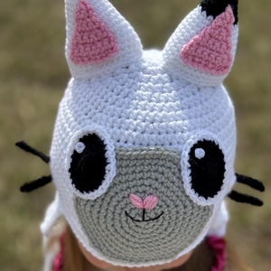 May include: A white crocheted cat hat with pink inner ears, black eyes, and black whiskers. The cat face is grey with a pink nose and a smiling mouth. The hat is handmade and designed to be worn.