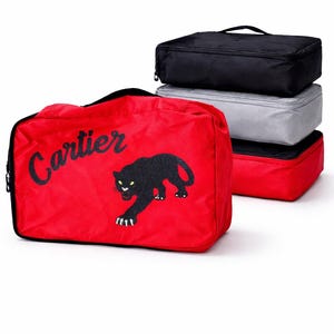 May include: Red travel bag with the word "Cartier" in black script and a black panther graphic. Stacked with three other travel bags in black, gray, and red. The bags are rectangular with zippers and handles.