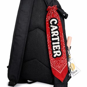 May include: Black backpack with a mesh back panel. A red bandana with a white paisley pattern and the word "CARTIER" in white is attached. A small tag that says "SQUAD" is attached to the backpack.