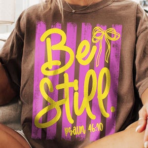 May include: Brown t-shirt with a vertical striped design in pink and purple. The shirt features the words "Be Still" in yellow script, with a yellow bow graphic and the text "psalm 46:10".