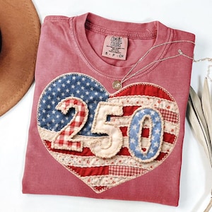 Puede incluir: Una camiseta Comfort Colors rosa polvoriento con un aplique en forma de corazón. El aplique presenta el número "250" en un diseño patriótico con telas rojas, blancas y azules. Un collar de plata con un pequeño colgante descansa sobre la camisa.