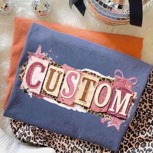 May include: A blue t-shirt with the word "CUSTOM" in a decorative font, layered over an orange shirt. The design includes pink, brown, and leopard print elements. A pair of leopard print pants and a white boot are also visible.
