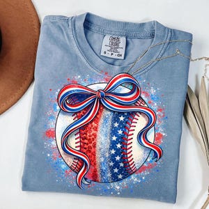 Könnte beinhalten: Hellblaues T-Shirt mit einem Baseball-Design mit roten, weißen und blauen Akzenten. Der Baseball hat eine Schleife und ein Design, das an die amerikanische Flagge erinnert. Das Shirt besteht aus einem weichen Material und trägt den Text "Comfort Colors" auf dem Etikett.
