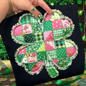 May include: A patchwork shamrock design in green, pink, and white fabrics, with a ruffled edge and gold shamrock accents. The word "LOVE" is stitched on the shamrock. The shamrock is on a black background.