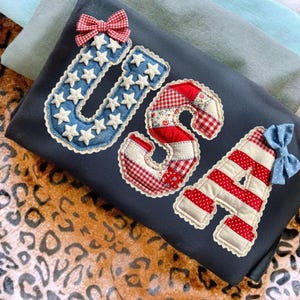 Può includere: Tessuto blu navy scuro con le lettere "USA" applicate in tessuto rosso, bianco e blu. La "U" è blu con stelle bianche e un fiocco a quadretti rossi. La "S" è a quadretti rossi e bianchi e con stampa floreale. La "A" è a righe rosse e bianche con un fiocco a pois blu.