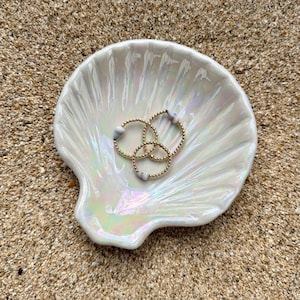 May include: A pearlescent white seashell-shaped dish holds three gold beaded rings, each with a small, light-colored stone. The dish sits on a bed of sand, creating a beachy, coastal aesthetic. The rings are a delicate jewelry item.