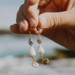 May include: Gold-toned earrings with a pearl and seashell design. Each earring features a teardrop-shaped white pearl, a smaller iridescent pearl, and a small seashell charm. The earrings are held up by a hand.