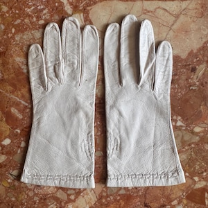 May include: A pair of off-white leather gloves featuring a textured, woven-style stitched pattern along the wrist cuffs and a small decorative stitched detail on the back of each hand, showcasing a soft, natural grain finish on the leather surface.