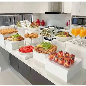 Könnte beinhalten: Ein weißer Buffettisch mit mehreren Ebenen, auf denen jeweils verschiedene Speisen wie Sandwiches, Obst und Gebäck präsentiert werden. Der Tisch steht in einer Küche mit weißen Schränken und einer Marmorarbeitsplatte.