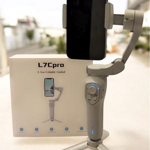 Op de afbeelding: Een grijze L7Cpro 3-assige opvouwbare gimbal met een smartphone bevestigd. Het apparaat staat op een statief en heeft bedieningsknoppen. De productdoos is wit met de productnaam en een afbeelding van de gimbal.