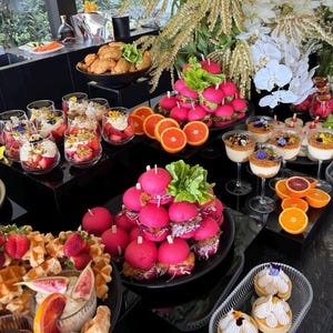 May include: A table laden with a selection of food and desserts, including pink cake pops, mini cheesecakes, and fruit. There are also orange slices and white flowers on the table.