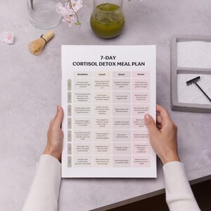Pode incluir: Uma pessoa segurando um plano de refei&ccedil;&otilde;es impresso de desintoxica&ccedil;&atilde;o de cortisol de 7 dias. O plano inclui op&ccedil;&otilde;es para caf&eacute; da manh&atilde;, almo&ccedil;o, lanche e jantar. O fundo inclui um copo de suco verde, um batedor de matcha e flores decorativas.