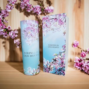 Könnte beinhalten: Eine blaue und weiße zylindrische Urne mit einem Blumendesign und dem Text "Forever in our hearts". Die Urne steht neben einer passenden Schachtel mit dem gleichen Design.