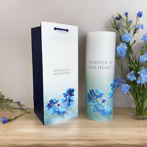 Könnte beinhalten: Eine weiße zylindrische Urne mit einem blauen Blumenmuster und dem Text "Forever in Our Hearts". Die Urne steht neben einer weißen Geschenktüte mit einem blauen Blumenmuster und dem gleichen Text.