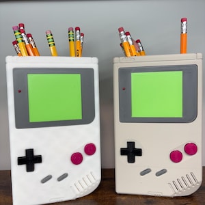 May include: Two pencil holders shaped like retro handheld game consoles. One is white, the other is beige. Each has a green screen, gray accents, a black cross-shaped D-pad, and pink buttons. Pencils with erasers are in the holders.