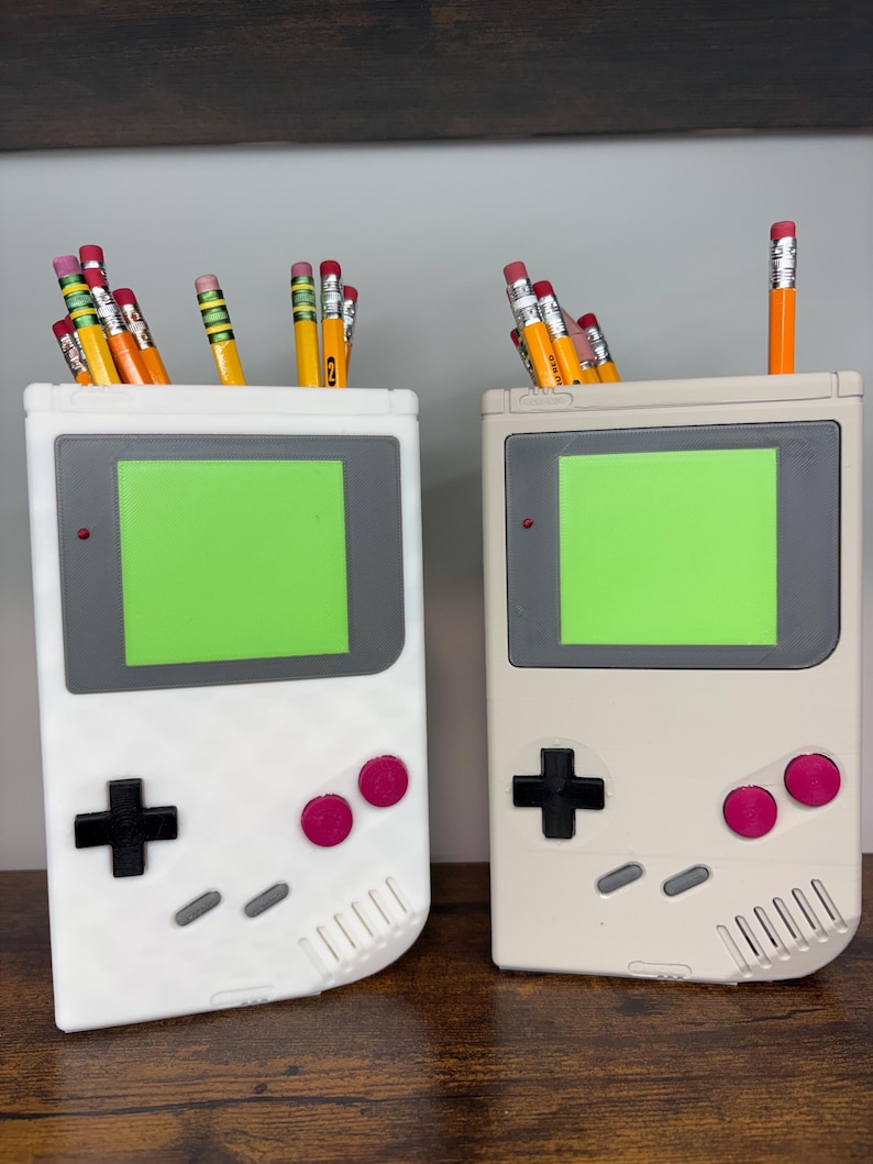 May include: Two pencil holders shaped like retro handheld game consoles. One is white, the other is beige. Each has a green screen, gray accents, a black cross-shaped D-pad, and pink buttons. Pencils with erasers are in the holders.