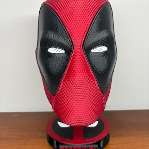 May include: A red and black textured Deadpool mask mounted on a circular stand. The mask features white mesh eye lenses and a smooth black eye area. The base is red and black with a rectangular plaque displaying the text DEADPOOL in red letters.