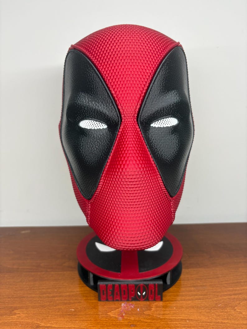 May include: A red and black textured Deadpool mask mounted on a circular stand. The mask features white mesh eye lenses and a smooth black eye area. The base is red and black with a rectangular plaque displaying the text DEADPOOL in red letters.