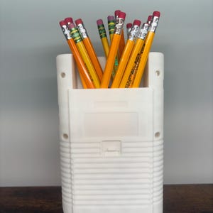 May include: A white pencil holder shaped like a retro handheld game console, filled with yellow pencils with pink erasers. The pencils are arranged upright, creating a functional and nostalgic desk accessory.