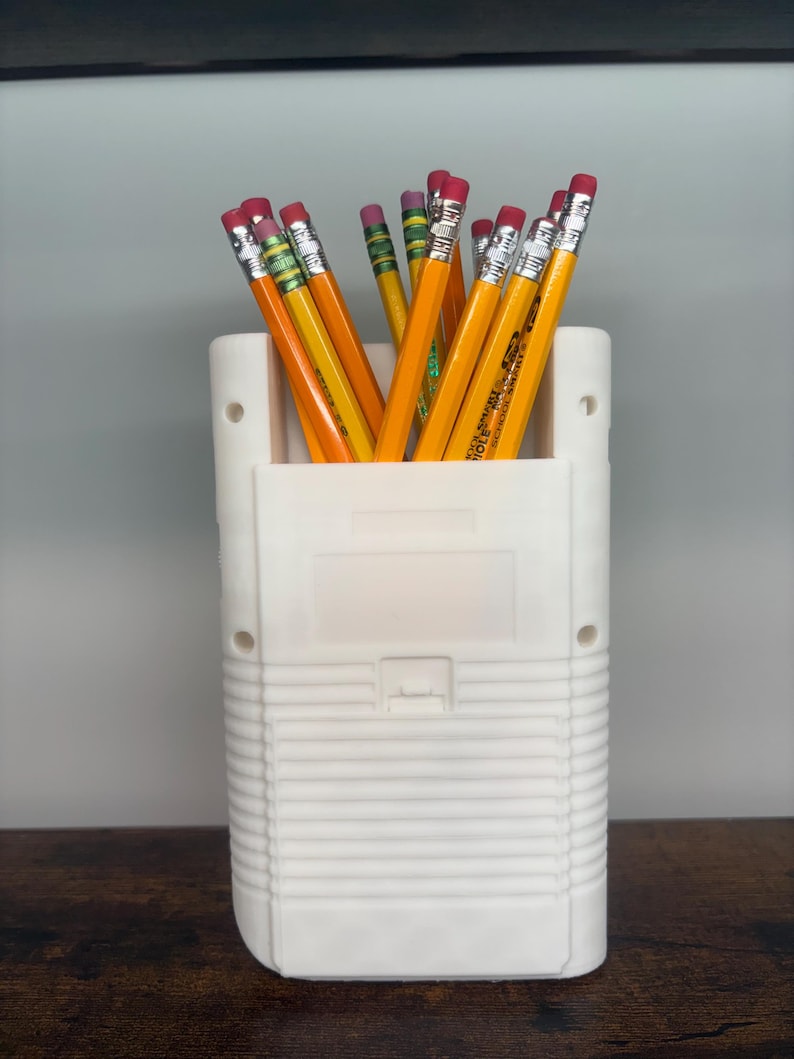 May include: A white pencil holder shaped like a retro handheld game console, filled with yellow pencils with pink erasers. The pencils are arranged upright, creating a functional and nostalgic desk accessory.