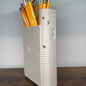 May include: A light gray pencil holder filled with yellow pencils with pink erasers. The pencil holder is rectangular with a textured design and a few circular holes. The pencils are arranged upright, creating a functional and organized desk accessory.