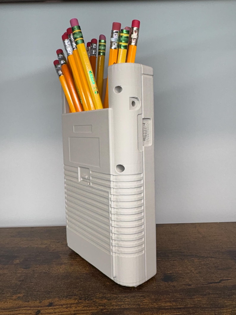 May include: A light gray pencil holder filled with yellow pencils with pink erasers. The pencil holder is rectangular with a textured design and a few circular holes. The pencils are arranged upright, creating a functional and organized desk accessory.