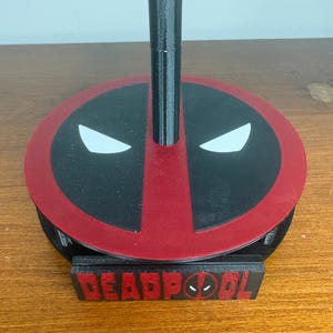 May include: A black and red circular stand featuring the Deadpool mask design with white eyes on the top surface. A vertical black pole extends from the center of the base. A rectangular black plaque with the red text DEADPOOL is attached to the front.