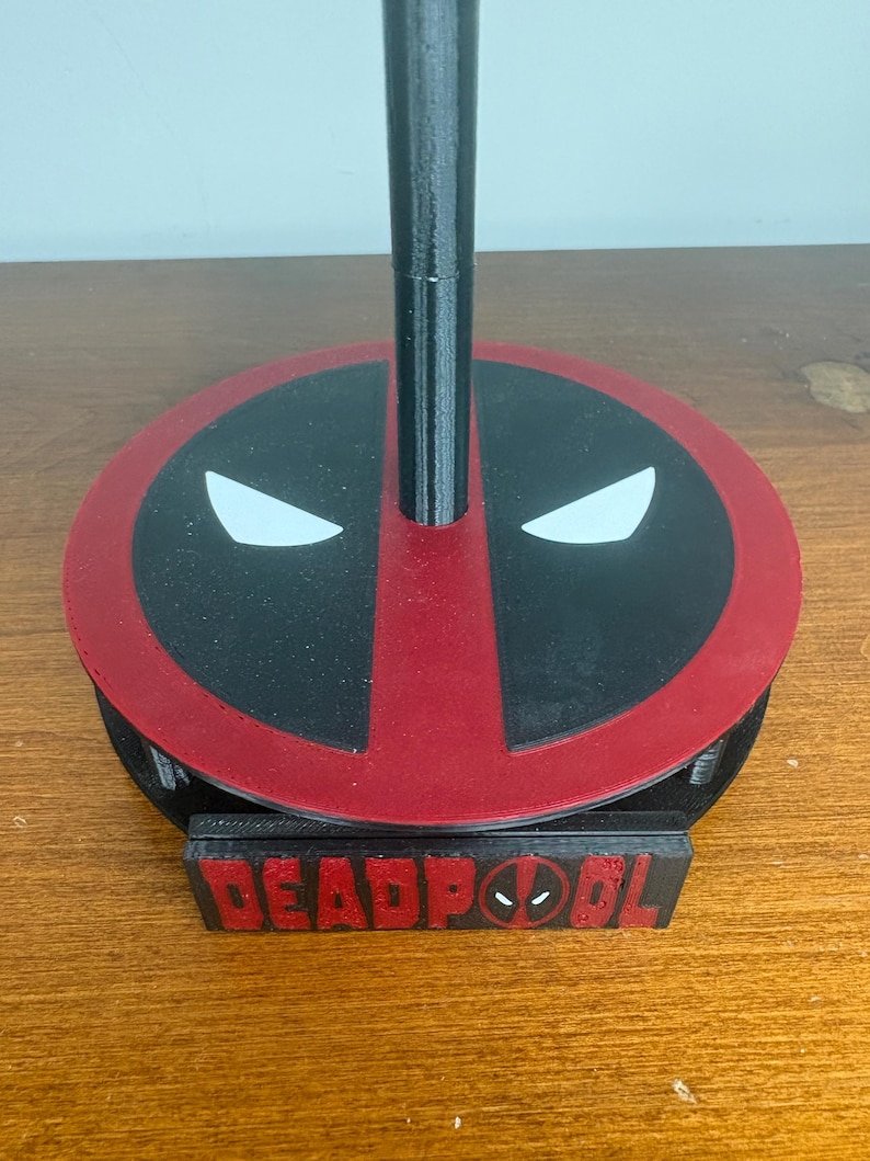 May include: A black and red circular stand featuring the Deadpool mask design with white eyes on the top surface. A vertical black pole extends from the center of the base. A rectangular black plaque with the red text DEADPOOL is attached to the front.