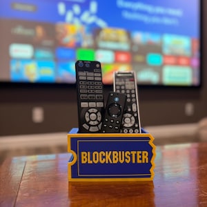 May include: A blue and yellow rectangular remote control holder shaped like a movie ticket with the word BLOCKBUSTER printed in yellow on the front. Three black electronic remote controls are standing upright inside the holder.