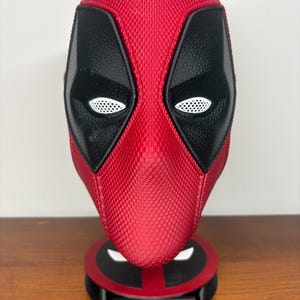 May include: A red and black textured mask featuring white mesh eye lenses, mounted on a circular stand with a base plate displaying the text DEADPOOL in red lettering. The mask has a distinct grid-like pattern across the red and black surfaces.