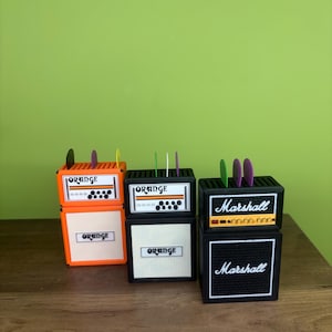 May include: Miniature orange and black amplifier replicas with the brand name "Orange" and "Marshall" displayed. Each amp has a speaker and colorful picks inserted into the top. The amps are displayed on a wooden surface against a green background.