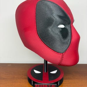 May include: A red and black textured Deadpool mask mounted on a circular display stand. The stand features a black and red base with white eye accents and a rectangular plaque at the front with the word DEADPOOL printed in red lettering.