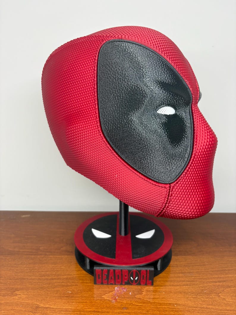 May include: A red and black textured Deadpool mask mounted on a circular display stand. The stand features a black and red base with white eye accents and a rectangular plaque at the front with the word DEADPOOL printed in red lettering.