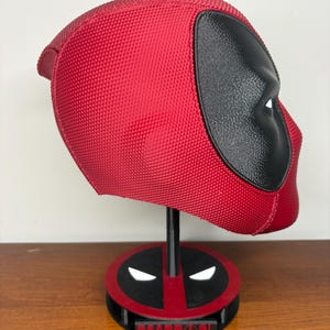 May include: A red and black textured Deadpool helmet displayed on a matching circular stand. The helmet features a prominent black eye patch area. The base of the stand is red and black with white eye accents and a black plaque with the text Deadpool.